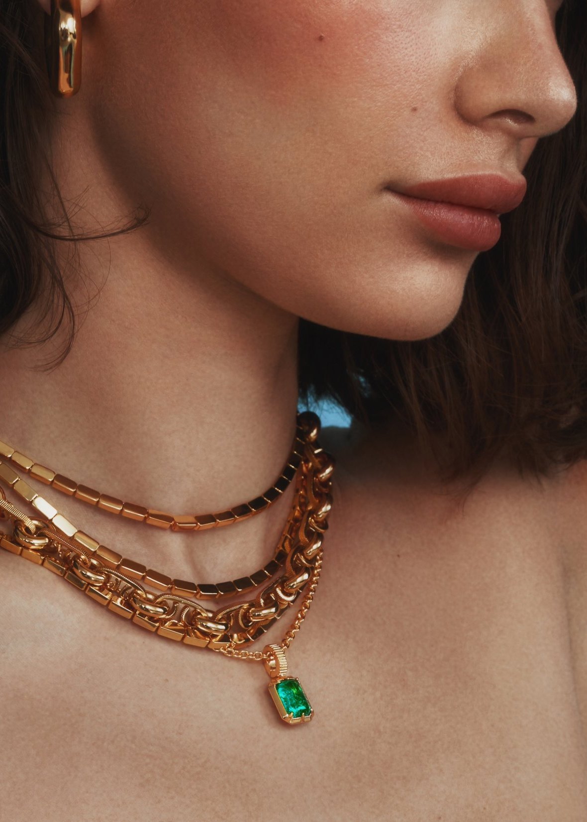 Close-up of the Kathryn Emerald Pendant layered with bold gold chains against a blue backdrop